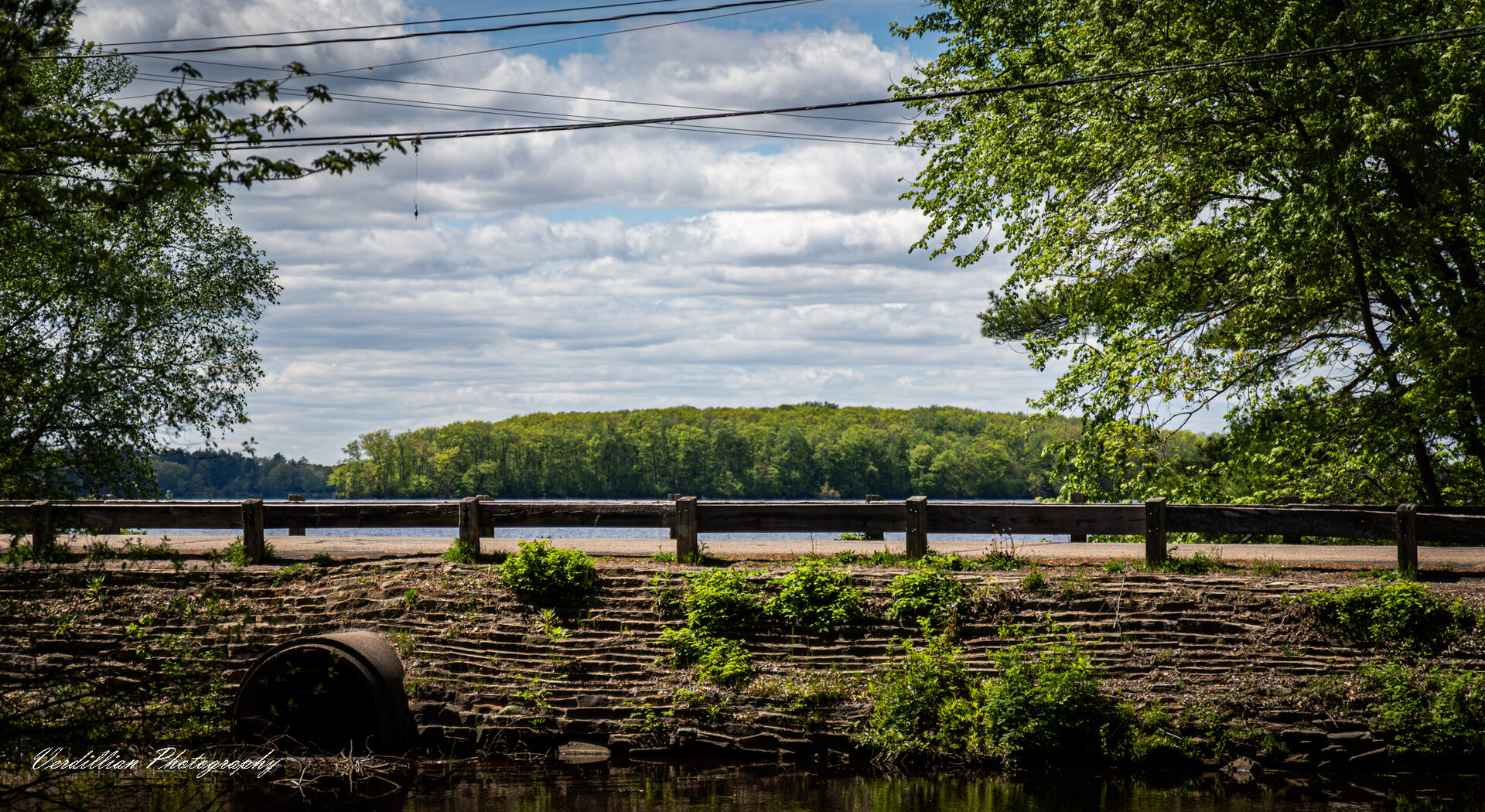 A wooden guardrail on a stone bridge overlooking a lake and treeline